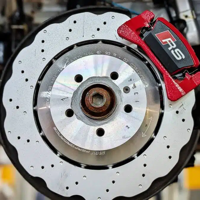 Audi VW Service Close-up of a ventilated and drilled car brake disc with a red brake caliper labeled RS. The metal disc shows wear, while the caliper is clean and prominent, indicating a high-performance braking system. Reflex Tuning, Hookett NH