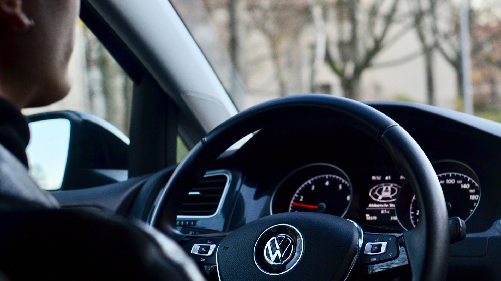 Audi VW Service A person sits in the drivers seat of a Volkswagen car, with a clear view of the steering wheel, dashboard, and speedometer. Trees and buildings are slightly blurred outside the window. Reflex Tuning, Hookett NH