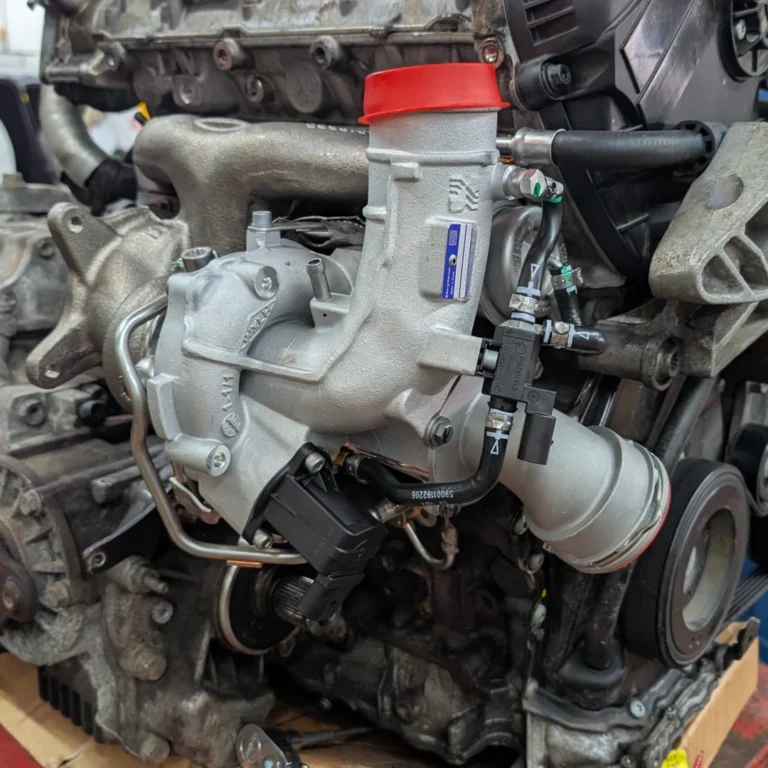 Audi VW Service A close-up view of a car engine with a turbocharger installed, featuring metallic components, hoses, and a red cap on the intake. The engine sits on a wooden surface in a workshop setting. Reflex Tuning, Hookett NH