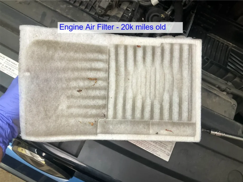 Audi VW Service A close-up of a used, dirty engine air filter labeled Engine Air Filter - 20k miles old, being held above a car engine by a gloved hand. The filter shows visible dust, debris, and discoloration. Reflex Tuning, Hookett NH