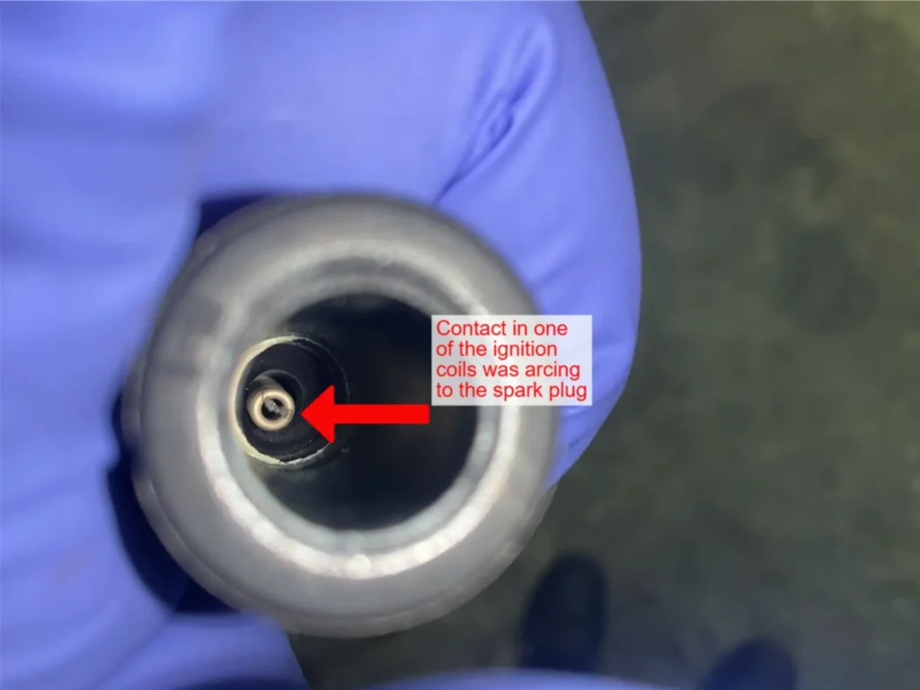 Audi VW Service A gloved hand holds an ignition coil, showing a close-up of the metal contact inside. A red arrow points to the contact with a label reading, “Contact in one of the ignition coils was arcing to the spark plug.”. Reflex Tuning, Hookett NH