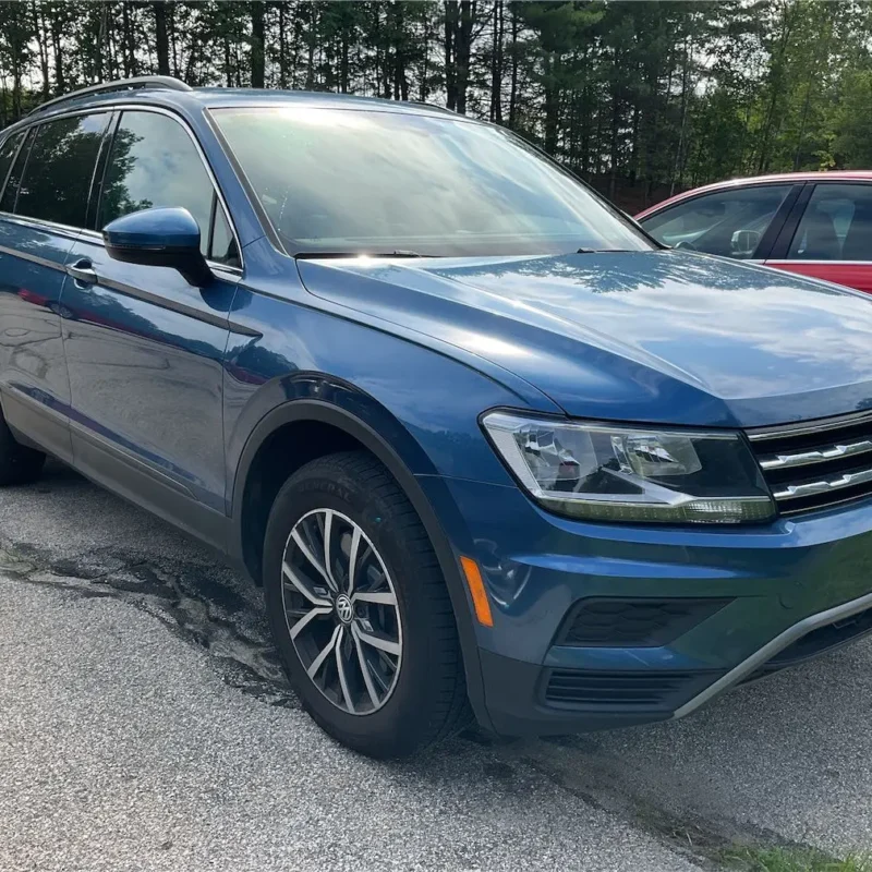 Audi VW Service A blue Volkswagen SUV is parked in a lot next to a red car, with trees in the background on a sunny day. Reflex Tuning, Hookett NH