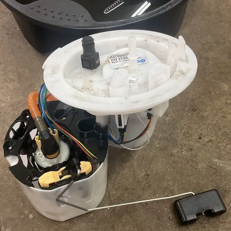 Audi VW Service A disassembled automotive fuel pump module with attached fuel lines, electrical wires, and a fuel level sensor is placed on a concrete floor near a black plastic container. Reflex Tuning, Hookett NH