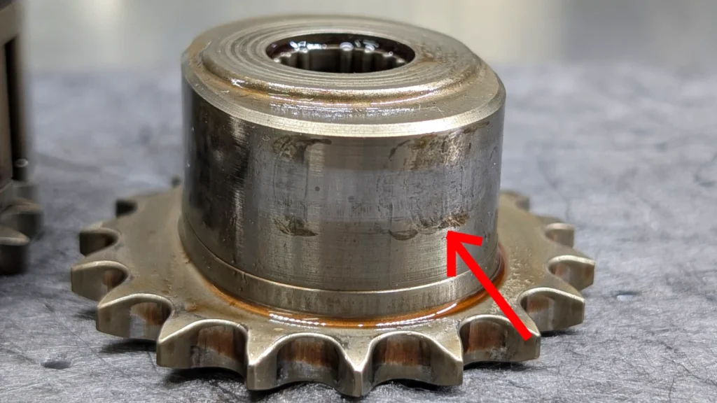 Audi VW Service A close-up of a metal sprocket with visible wear marks on its surface. A red arrow points to the worn area, highlighting scratches and discoloration on the gears cylindrical body. Reflex Tuning, Hookett NH