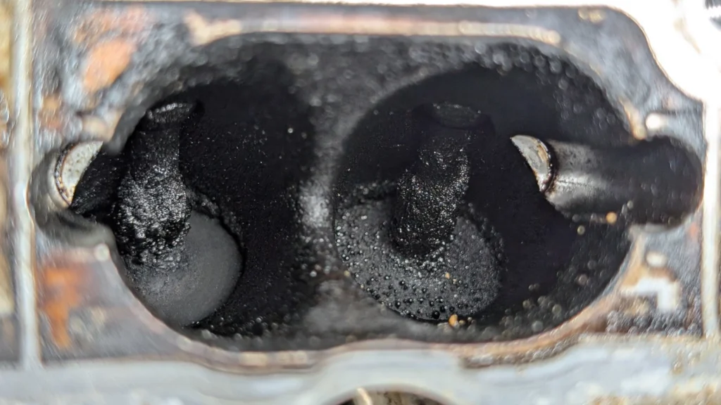 Audi VW Service Close-up view of two engine intake valves heavily coated with black carbon deposits, indicating buildup and possible engine performance issues. The surrounding metal shows signs of wear and oxidation. Reflex Tuning, Hookett NH