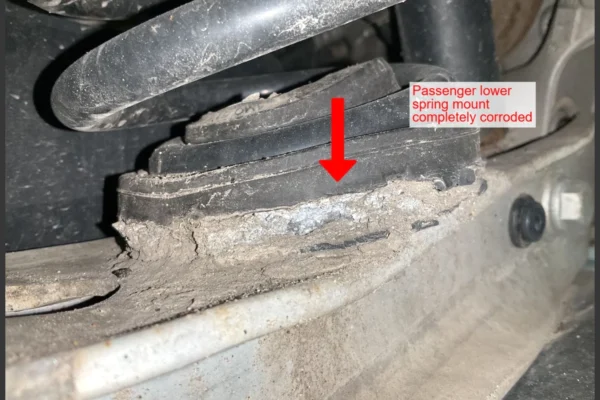 Audi VW Service Close-up of a car suspension component with visible rust and corrosion on the passenger lower spring mount. A red arrow points to the damaged area, with a label noting it is completely corroded. Reflex Tuning, Hookett NH