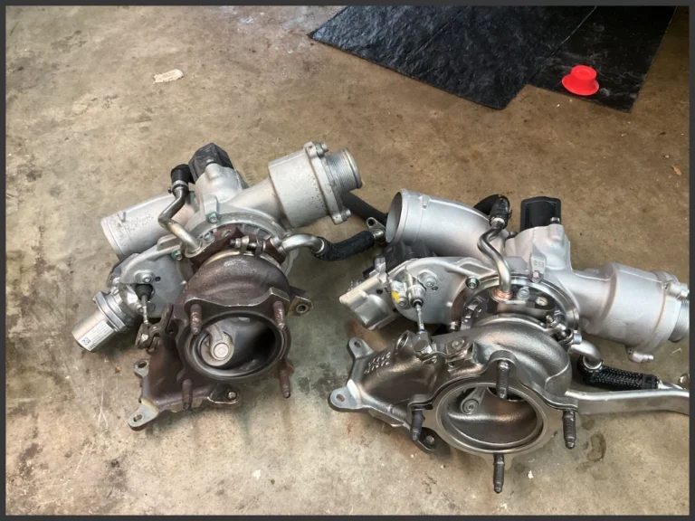 Audi VW Service Two turbochargers sit side by side on a concrete floor, one appearing old and worn with darker coloration, and the other looking new and clean with shiny metal surfaces and attached components. Reflex Tuning, Hookett NH