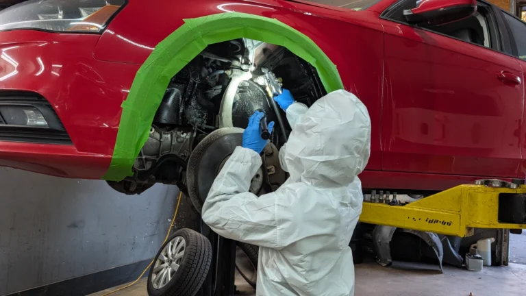 Audi VW Service A person in protective gear and blue gloves works on the wheel area of a red car lifted on a hydraulic lift, with green tape around the wheel arch. Reflex Tuning, Hookett NH