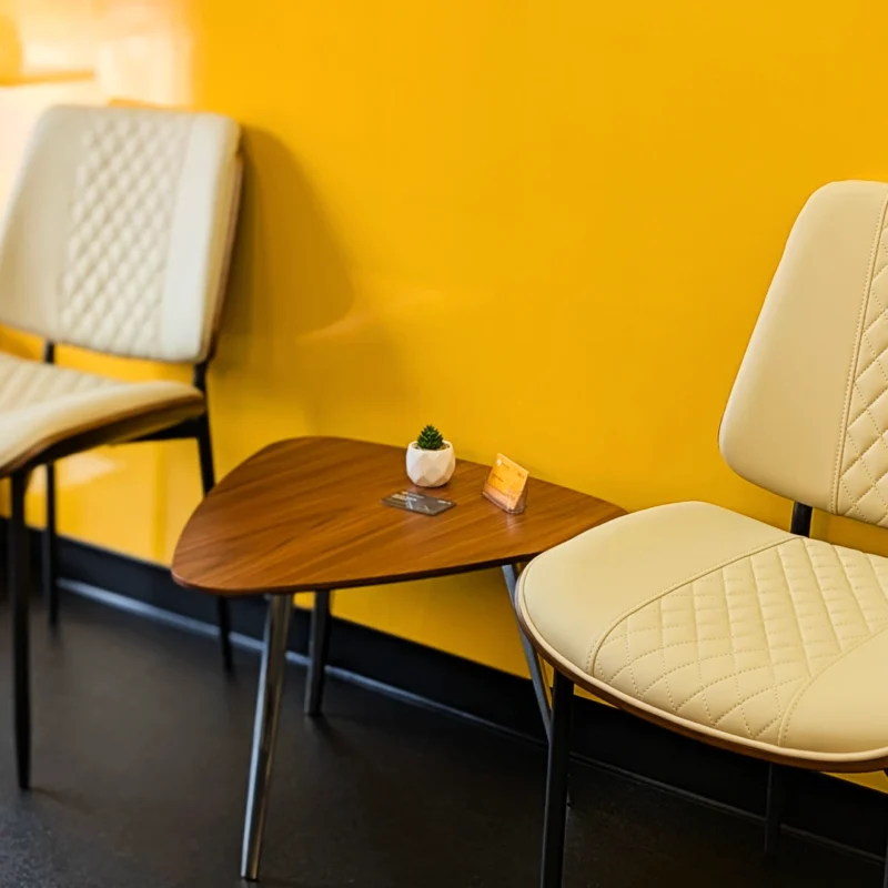 Audi VW Service Two cream-colored chairs with quilted patterns sit next to a small, triangular wooden table holding a tiny plant and card, against a bright yellow wall and black floor. Reflex Tuning, Hookett NH