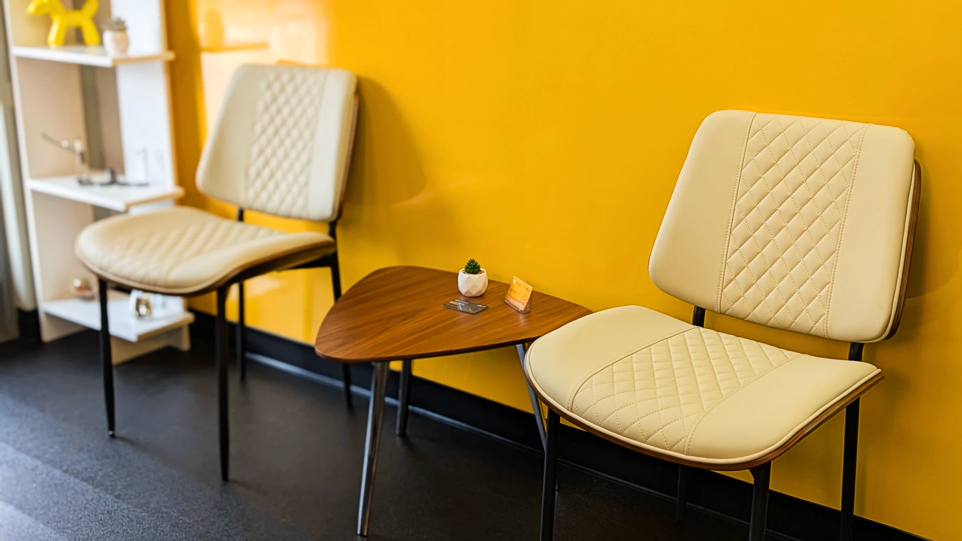 Audi VW Service Two cream-colored chairs with quilted patterns sit next to a small, triangular wooden table holding a tiny plant and card, against a bright yellow wall and black floor. Reflex Tuning, Hookett NH
