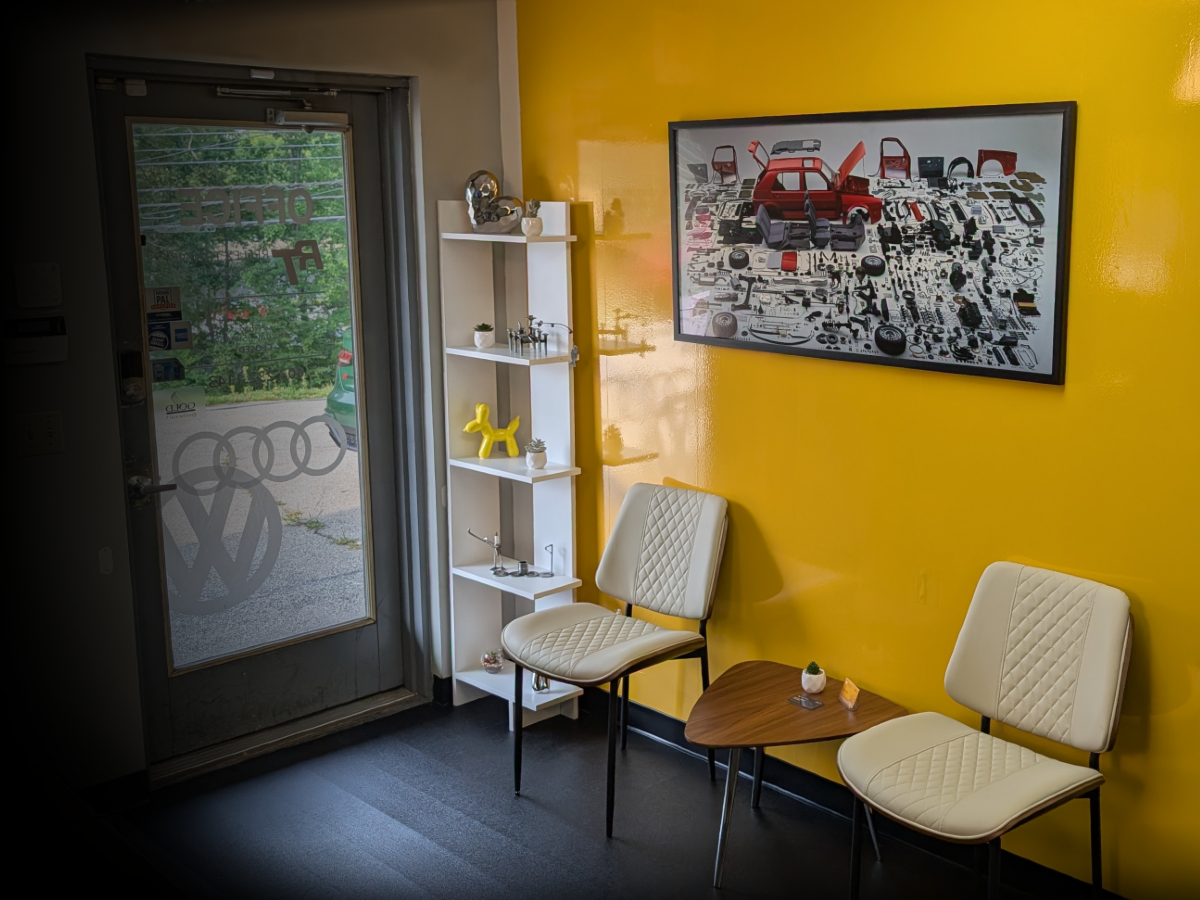 Audi VW Service A waiting area with two cream chairs and a small table against a bright yellow wall, decorated with a framed car parts poster and a white shelving unit; a glass door with car logos is on the left. Reflex Tuning, Hookett NH