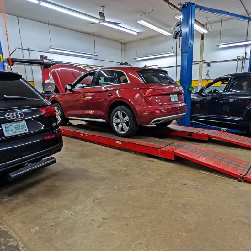 Audi VW Service Three SUVs, two black and one red, are parked inside an auto repair shop. The red SUV is on a car lift ramp, and the shop has bright overhead lights and various equipment visible. Reflex Tuning, Hookett NH