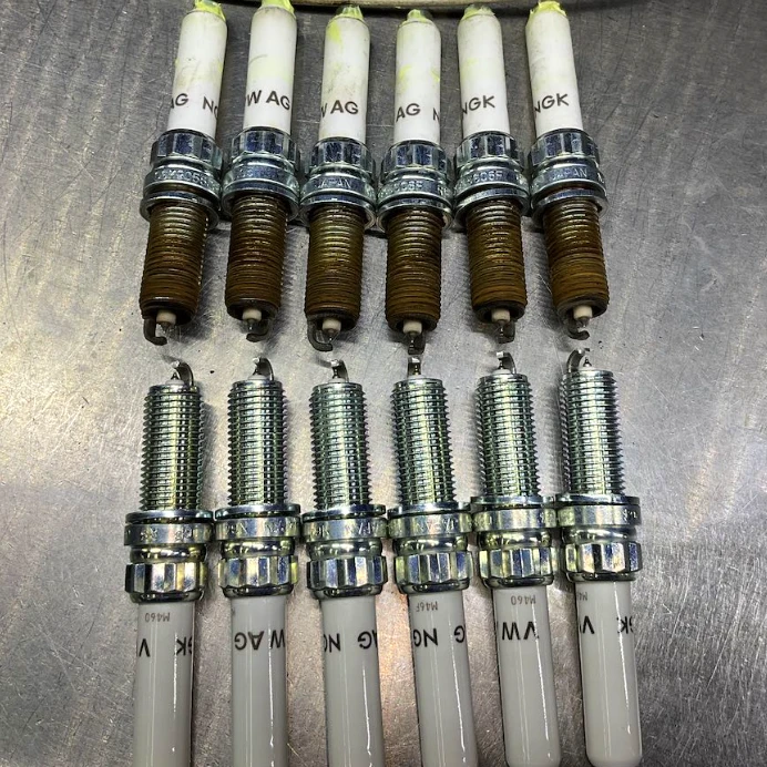 Audi VW Service Ten spark plugs arranged in two rows on a metallic surface; the top row shows five used, dirty spark plugs, while the bottom row displays five clean, new spark plugs. Reflex Tuning, Hookett NH
