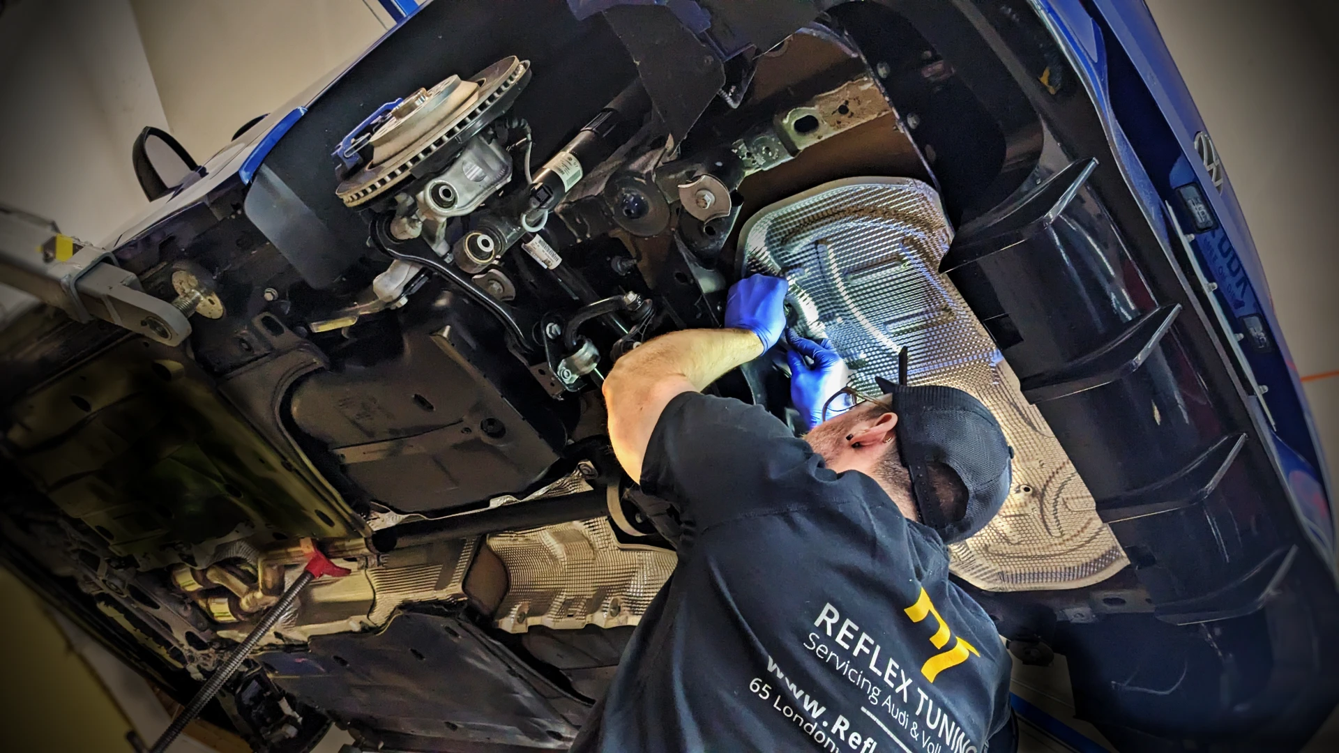Audi VW Service A mechanic wearing gloves and a cap works underneath a raised car, using tools to repair or inspect parts near the exhaust system in an auto service garage. Reflex Tuning, Hookett NH